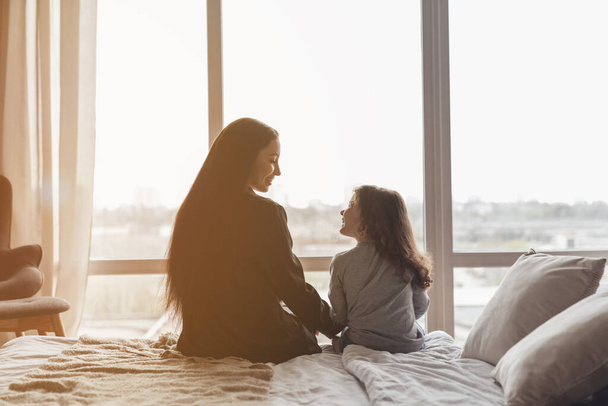 Mom and daughter woke up in the morning sitting on the bed in front of a large window. - Foto, afbeelding