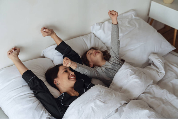 Mom and daughter waking up after sleeping in their bed together and stretching their arms up in the bedroom at home. - Photo, Image