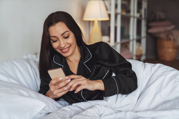 Egy fiatal, gyönyörű nő most ébredt fel az ágyban alvás után, és mosollyal a pizsamájában használja a telefont egy takaró alatt a hálószobájában.. - Fotó, kép