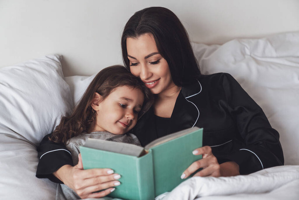 おやすみなさい。若い美しい母親は彼女の小さな娘をベッドに置き、彼女に夜の子供の物語の本を読む. - 写真・画像