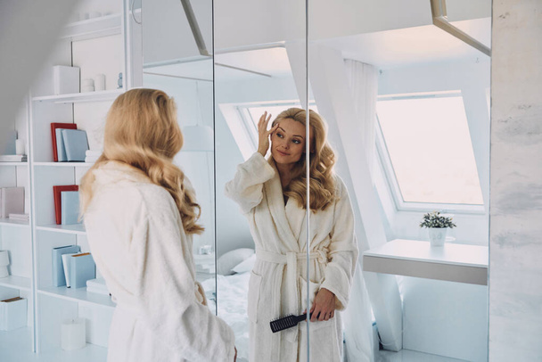 Attractive young woman in bathrobe looking at her reflection in the mirror - Fotoğraf, Görsel