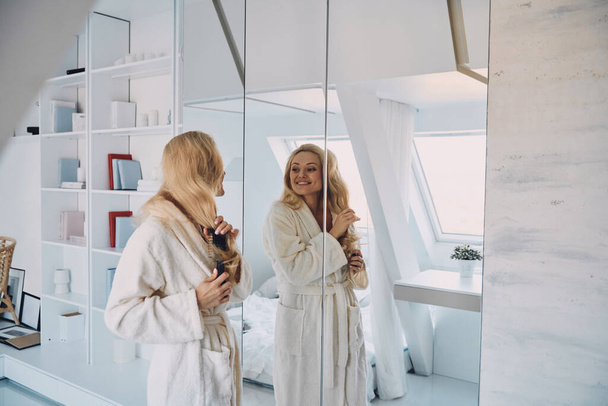 Приваблива молода усміхнена жінка в халаті розчісує волосся, стоячи перед дзеркалом вдома
 - Фото, зображення