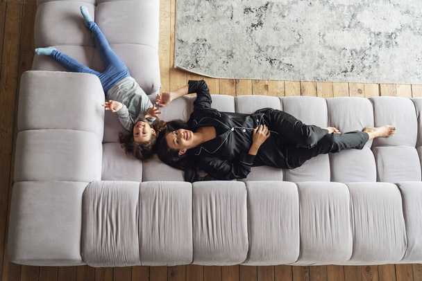 Young mother and her little daughter relax lying on the couch in the living room at home, smiling at the camera, top view. - Foto, imagen