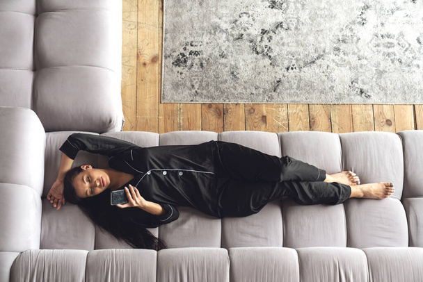 Молодая красивая женщина расслабляет лежа на диване в гостиной на дому, использует телефон, вид сверху. - Фото, изображение