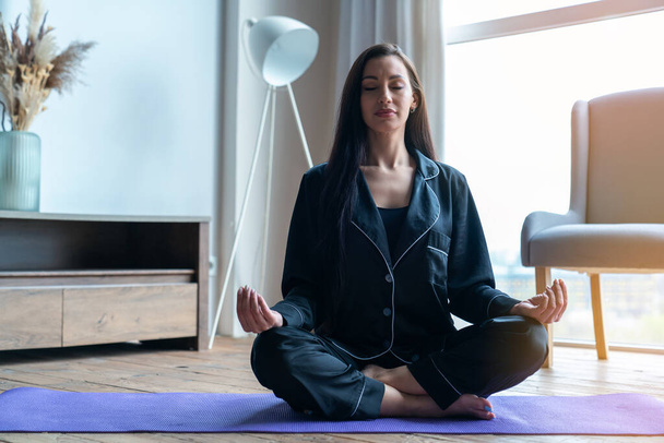 Junge schöne Fitness-Frau zu Hause sitzt in Lotusposition auf Matte beim Yoga-Kurs im heimischen Wohnzimmer. - Foto, Bild