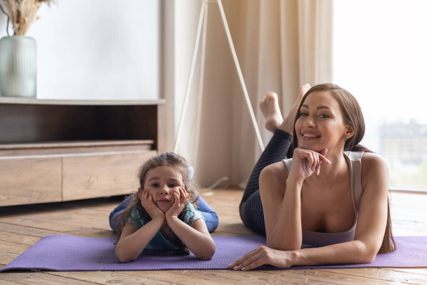 Młoda matka i jej córeczka leżące na macie na podłodze podczas zajęć fitness w salonie w domu i uśmiechnięte do kamery. - Zdjęcie, obraz