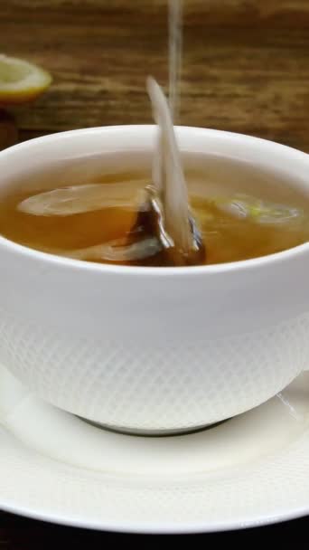 Sacchetto di tè preparato in tazza di ceramica bianca, immerso in acqua calda bollita e due pezzi di zucchero raffinato aggiunto. Il limone affettato giace sul tavolo di legno. Colpo verticale. - Filmati, video