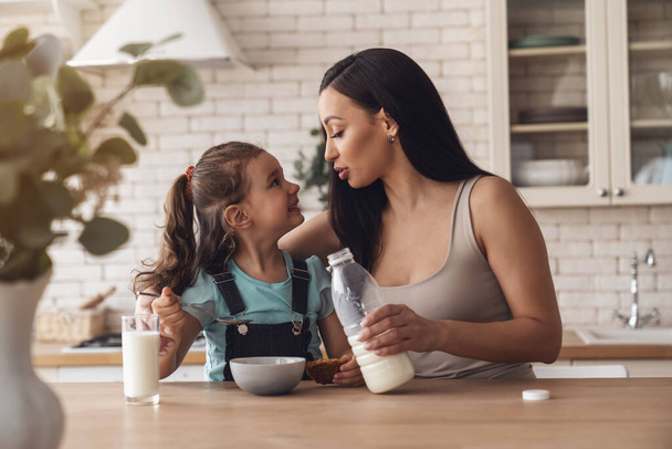 若いです幸せな母親準備朝食のために彼女の小さな娘によって追加ミルクとmuesliにプレートに座っている間に台所のテーブルに自宅. - 写真・画像