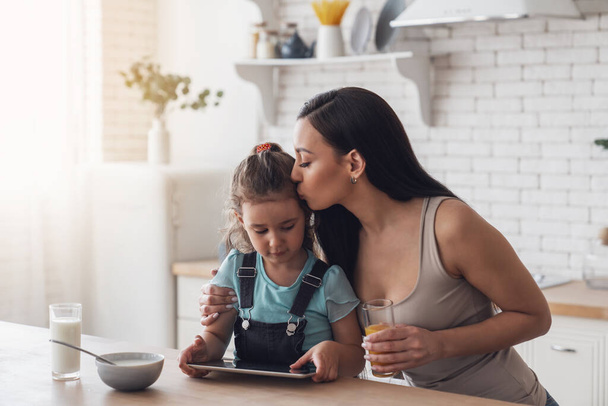Μια νεαρή ευτυχισμένη μητέρα ετοιμάζει πρωινό για τη μικρή της κόρη προσθέτοντας γάλα και μούσλι σε ένα πιάτο ενώ κάθεται σε ένα τραπέζι στην κουζίνα στο σπίτι και χρησιμοποιώντας ένα ηλεκτρονικό tablet. - Φωτογραφία, εικόνα