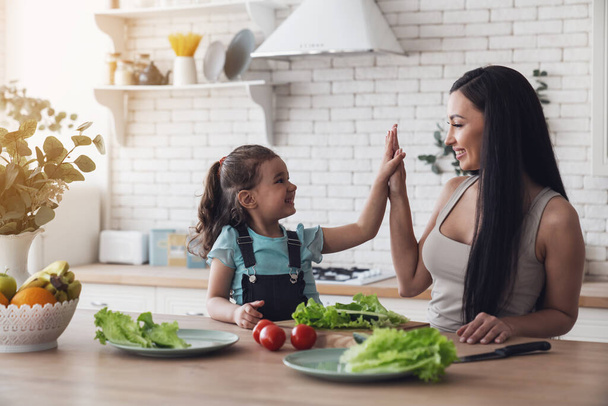 La piccola bambina felice aiuta sua madre a preparare un'insalata per loro a pranzo, seduta a un tavolo in cucina a casa.. - Foto, immagini