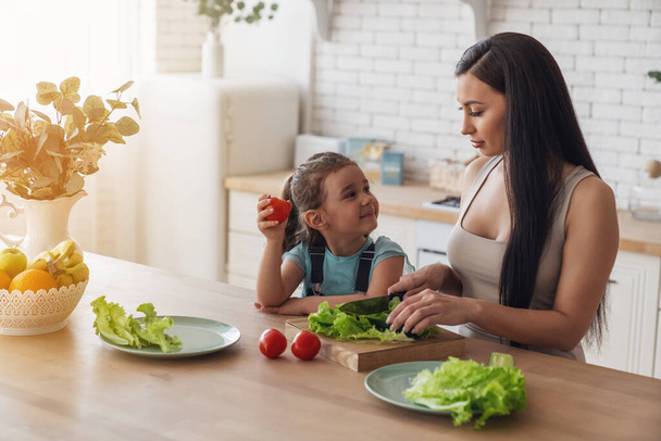 Little happy baby girl helps her mother to prepare a salad for them for lunch, sitting at a table in the kitchen at home. - Foto, imagen