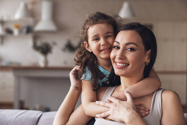 Happy young mother and her little daughter spend time together and have fun sitting hugging on the couch in the living room at home. - Photo, Image