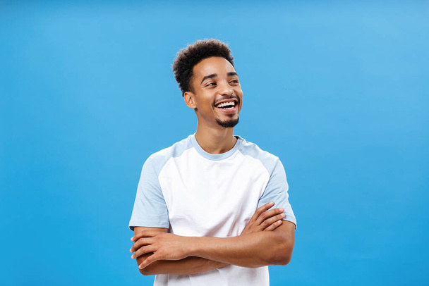 Proud and carefree cool african american male student in trendy t-shirt holding hands crossed on chest in self-assured carefree gesture turning right laughing pleased, having fun over blue wall - Photo, Image