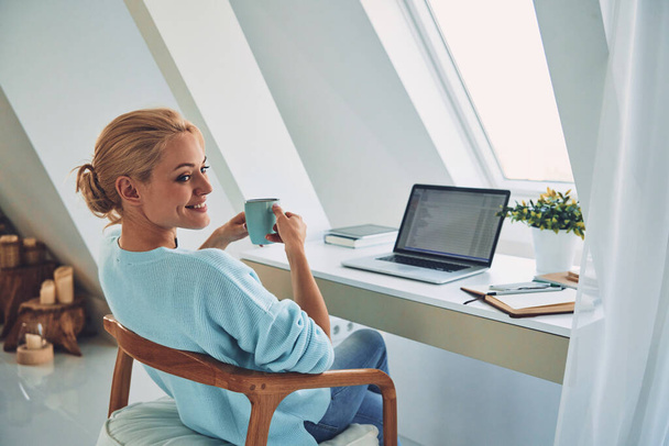 Rear view of young smiling woman enjoying hot drink while working at home - Foto, Bild