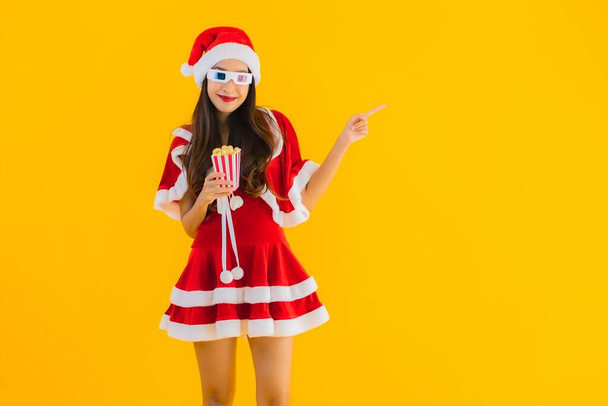 Portrait beautiful young asian woman wear christmas clothes hat with popcorn and 3d glasses ready for watch movie on yellow isolated background - Foto, imagen