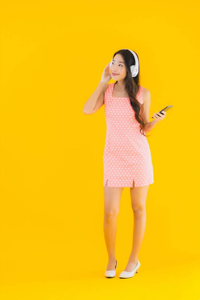 Портрет красивої молодої азіатки слухати музику зі смартфоном і навушниками на жовтому ізольованому фоні
 - Фото, зображення