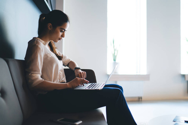 Side view of concentrated young ethnic female with ponytail in classy outfit sitting on couch and typing on laptop during work in modern office - Photo, Image