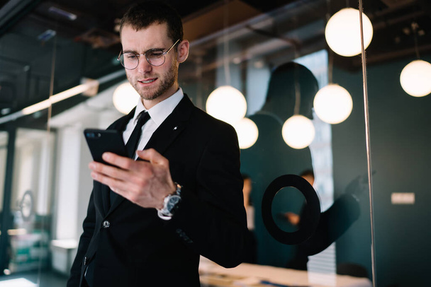 Crop positive businessman in black formal suit browsing modern mobile phone and smiling while standing near glass wall in contemporary business center - Foto, imagen