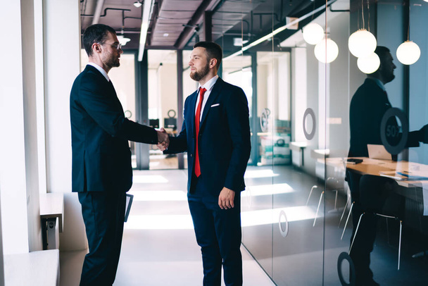 Serious male coworkers in formal clothes standing in modern office corridor together and greeting each other while discussing business project - Foto, Bild