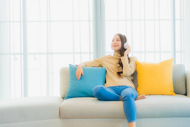 Portré gyönyörű fiatal ázsiai nő fejhallgatóval zenét hallgatni kanapén a nappaliban belső - Fotó, kép