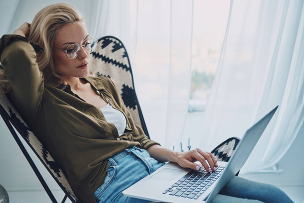 Attractive young woman using laptop while sitting in comfortable chair at home - Photo, image