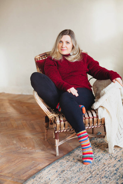 Attraente donna felice di mezza età seduta e rilassante sulla comoda sedia a casa il fine settimana, indossando calzini colorati a maglia e caldo maglione rosso, sorridendo alla macchina fotografica. Stile di vita persone - Foto, immagini