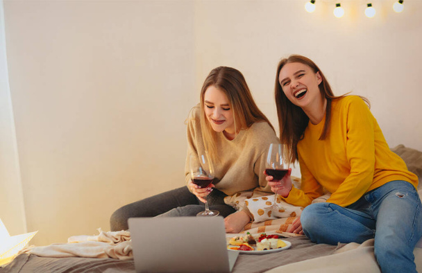 Twee leuke vrolijke grappige vriendinnen die plezier hebben samen, regelen home cinema avond, kijken comedy op laptop, lachen luid, zitten op bed met kaas bord en glazen rode wijn - Foto, afbeelding
