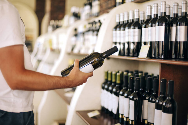 Anonimo cliente maschio esaminando bottiglia mentre in piedi vicino scaffali e selezionando il vino in negozio - Foto, immagini