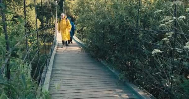Caminantes hombres y mujeres en impermeables caminan en puente colgante. Día despejado - Imágenes, Vídeo