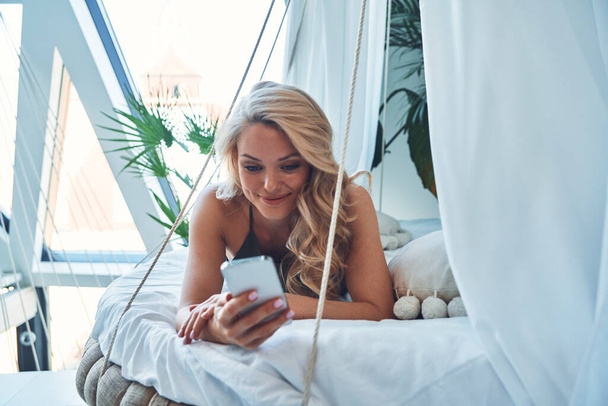 Beautiful young woman using smart phone and smiling while relaxing on bed at home - Foto, imagen