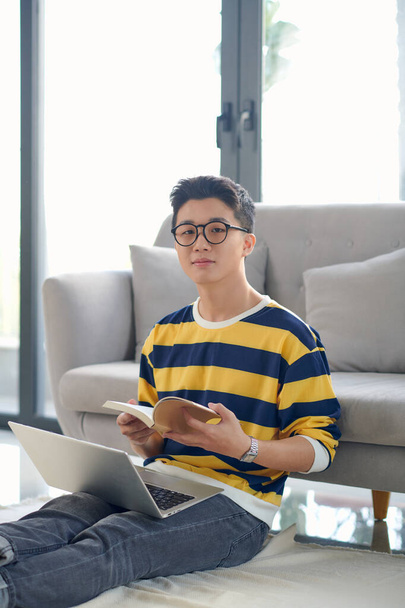 アジア人男性が床に座ってノートパソコンで本と仕事をしている - 写真・画像