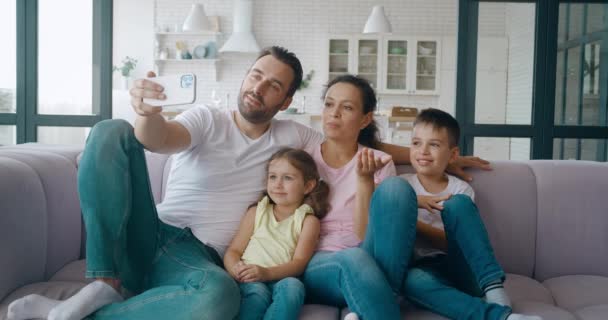 Volwassen vader, man zit naast zijn mooie vrouw en kinderen, houdt een mobiele telefoon vast en houdt een familievideoconferentie tijdens een videogesprek. Familie zwaaiende hand op zoek naar mobiele telefoon scherm - Video