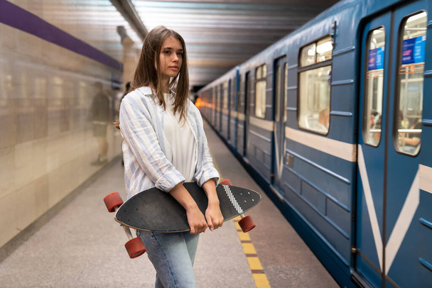 Chica joven en la plataforma del metro mira el coche del metro en la estación de metro subterráneo solo sosteniendo longboard - Foto, imagen