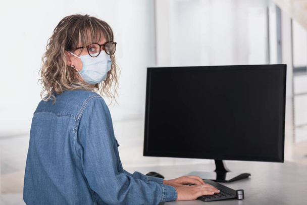Rückansicht einer erwachsenen Frau mit schützender Gesichtsmaske im Büro mittels Computer mit dunklem Bildschirm, um Raum zu kopieren - Foto, Bild