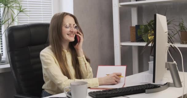 Cheerful girl secretary at her desk is talking on phone. Young business woman in office solves working issues. Nice phone conversation with a business partner - Footage, Video