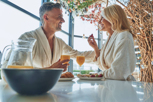 Beautiful mature couple in bathrobes enjoying breakfast together while spending time in the domestic kitchen - Fotoğraf, Görsel