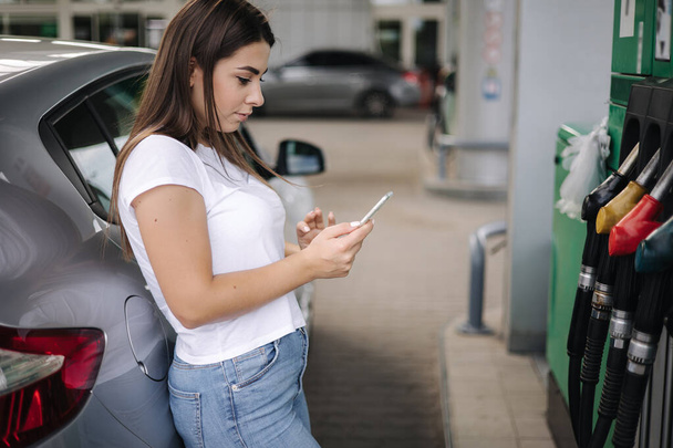 Uma jovem atraente usando seu telefone e apoiou-se em seu carro no posto de gasolina. Mulher espera posto de gasolina trabalhador - Foto, Imagem