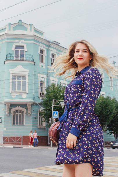 Young beautiful woman in blue dress with a round bag walking on the spring street. Urban background. Stylish Tourist girl enjoying walking the city during weekend trip. - Φωτογραφία, εικόνα