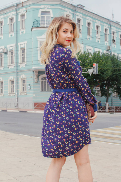Young beautiful woman in blue dress with a round bag walking on the spring street. Urban background. Stylish Tourist girl enjoying walking the city during weekend trip. - Φωτογραφία, εικόνα