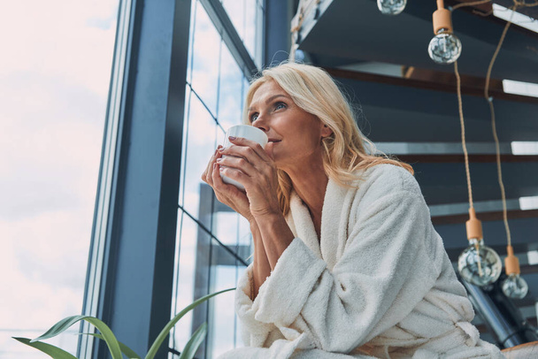 Beautiful mature woman in bathrobes drinking coffee and smiling while standing near window at home - Fotó, kép