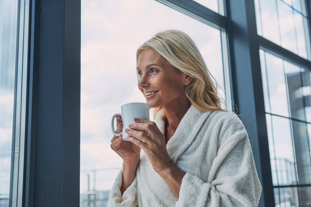Beautiful mature woman in bathrobes drinking coffee and smiling while standing near window at home - Foto, Bild