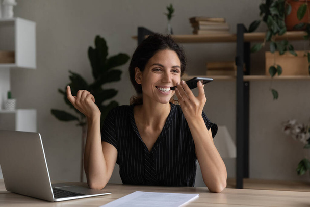 Smiling businesswoman recording voice message on smartphone, sitting at desk - Foto, Imagem
