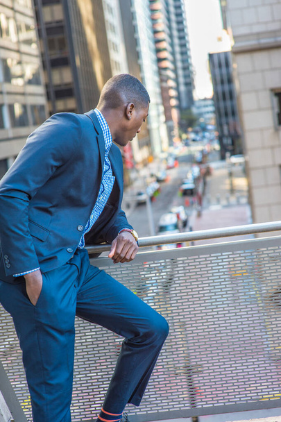 Ένας νεαρός μαύρος επιχειρηματίας στέκεται στο μπαλκόνι, σκύβοντας πάνω από τα κάγκελα, κοιτάζοντας κάτω στο δρόμο, χαλαρώνοντας - Φωτογραφία, εικόνα