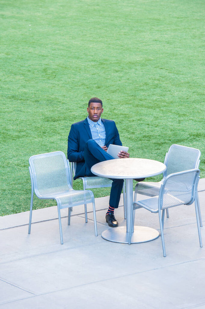 Молодий красивий чорний чоловік сидить на стільці біля зеленого газону і читає на маленькому комп'ютері
 - Фото, зображення