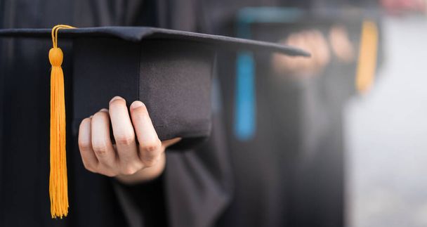 Zdjęcie Stockowe zamknięte i selektywne skupienie absolwentów uczelni wyższych w sukni dyplomowej gospodarstwa kapelusz dyplomowy w dniu rozpoczęcia - Zdjęcie, obraz