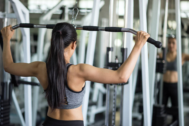 Rear fit young Asian woman doing exercise using machine in fitness gym. pumping back muscles. Bodybuilding and healthy lifestyle concept. Happy girl weight training in sport club with body reflection on mirror. - Foto, imagen