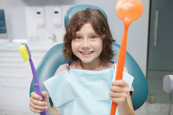 Muchacho joven emocionado sonriendo a la cámara, sosteniendo un gran espejo dental y cepillo de dientes en el consultorio de dentistas - Foto, Imagen