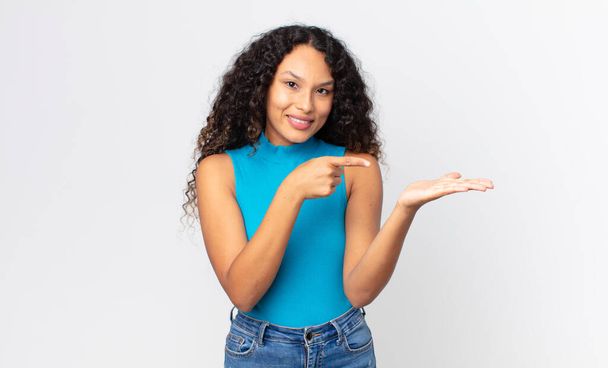 pretty hispanic woman smiling cheerfully and pointing to copy space on palm on the side, showing or advertising an object - 写真・画像
