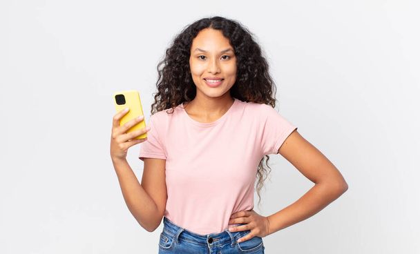 hispanic pretty woman smiling happily with a hand on hip and confident and holding a smartphone - Foto, Bild