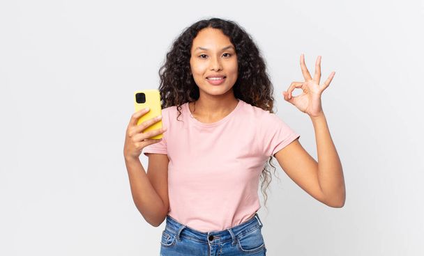 hispanic pretty woman feeling happy, showing approval with okay gesture and holding a smartphone - Photo, Image